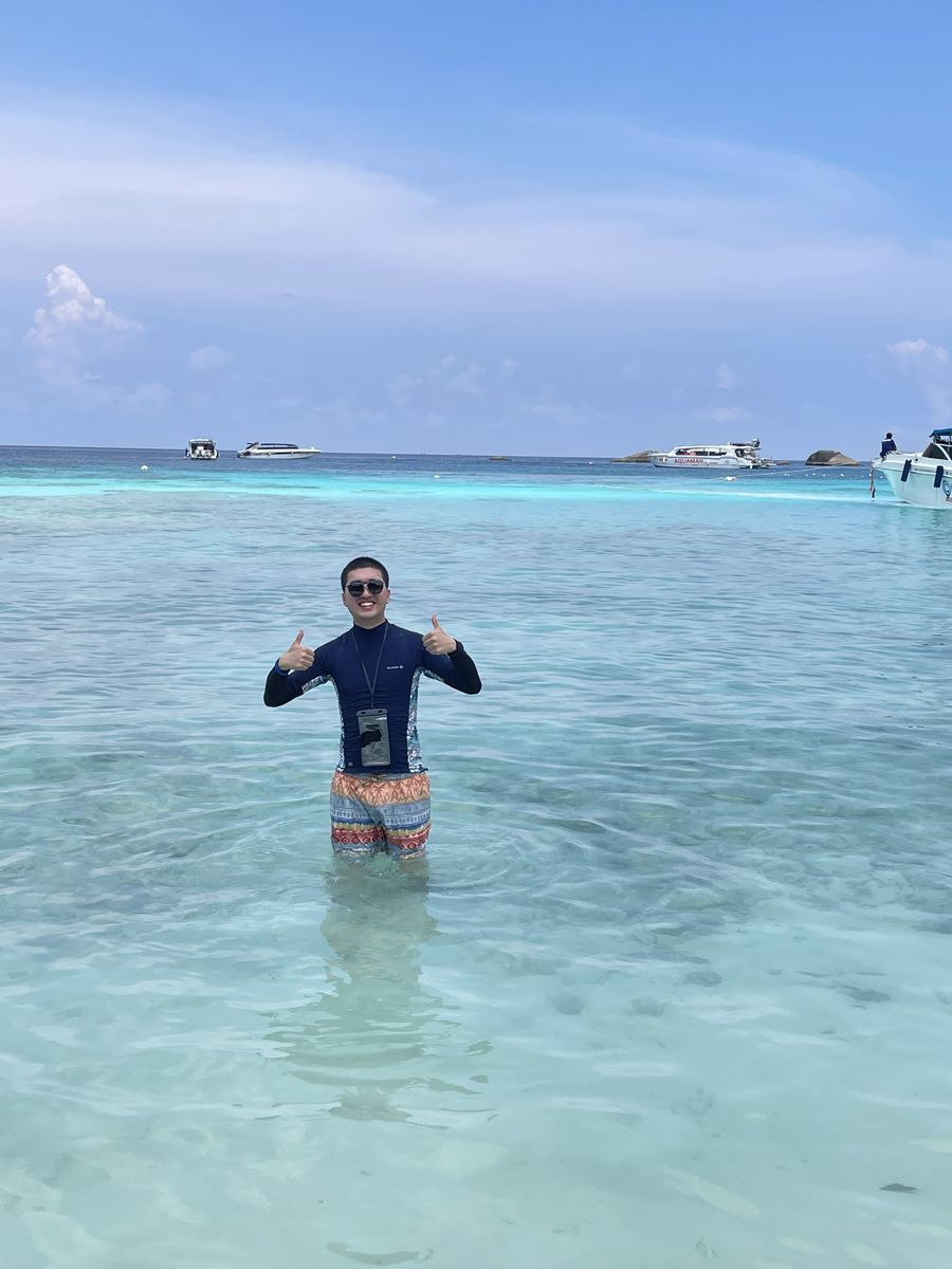 Tom standing waist-deep in clear turquoise water at the Similan Islands, Thailand, giving two thumbs up in a rash guard and sunglasses